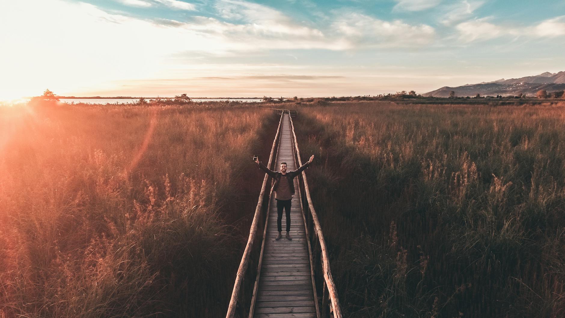 An Aerial Photography of a Man Standing on a Wooden Bridge with His Arms Open