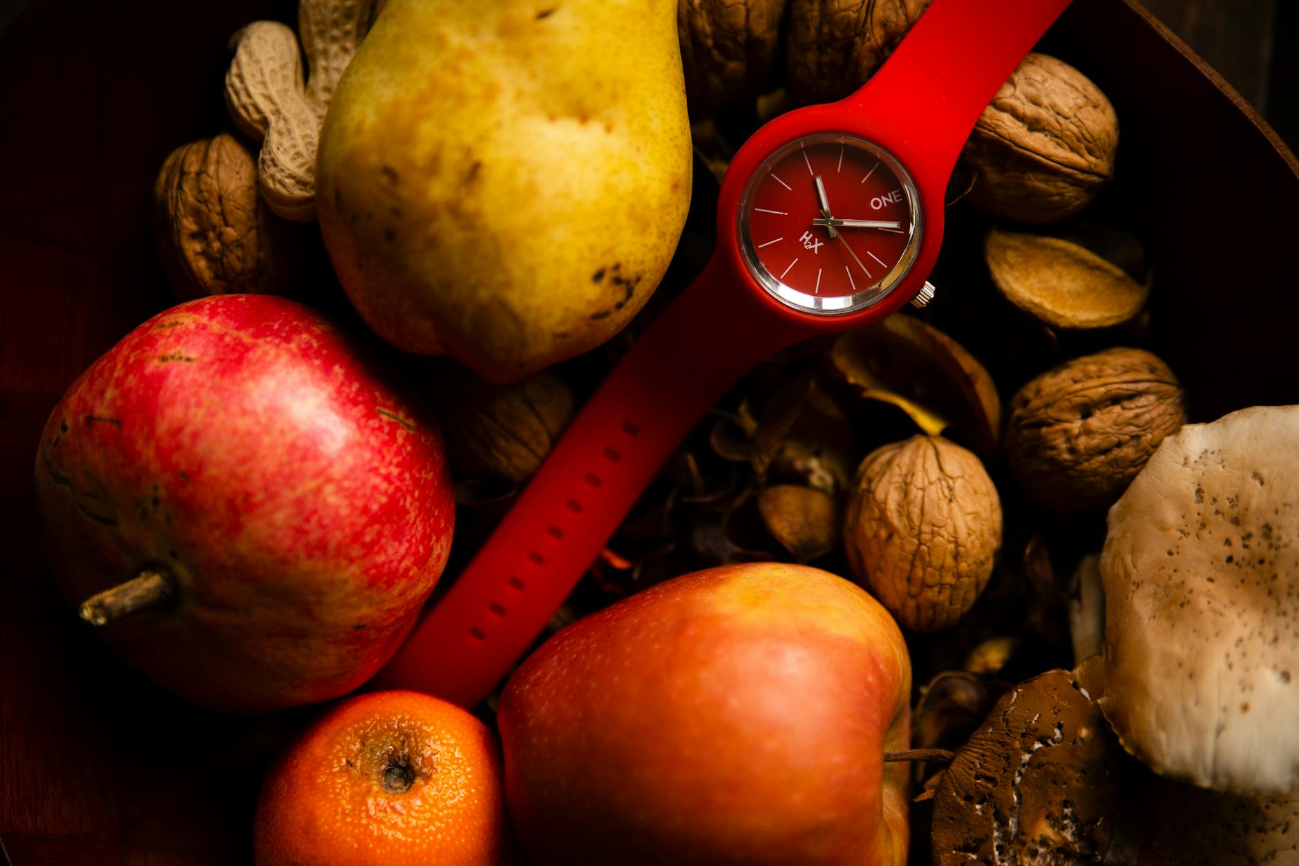 From above of red rubber wristwatch placed in healthy basket with organic fresh apples pear and orange and composed with different nuts